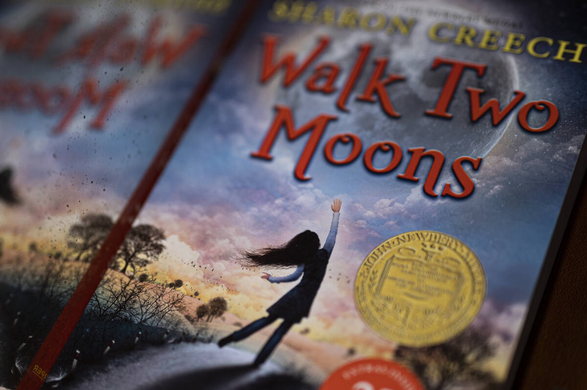 A copy of the book “Walk Two Moons” by Sharon Creech. Its cover depicts a girl reaching for a blue, pink, and purple sky as she stands on a road.