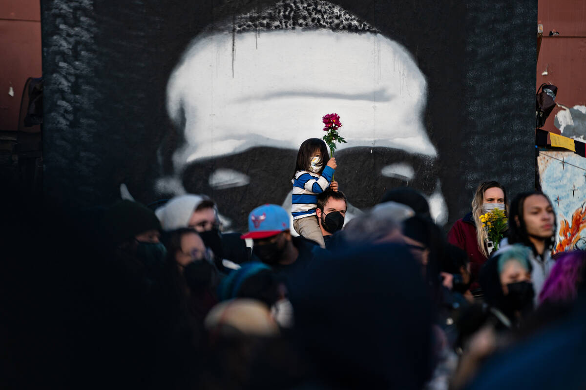 A child with long hair and a blue striped shirt sits on his father's shoulders while holding flowers. They are surrounded by other people celebrating in front of a mural of George Floyd.